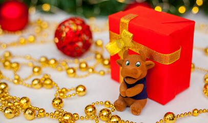 Toy brown bull near a red gift box on a white background. Christmas tree branch with red toys and golden garlands in the background. Bull as a symbol of the new year 2021. New Year, Christmas concept.