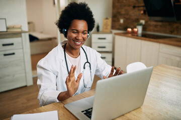 Happy black doctor waving while greeting someone during video call from her office.
