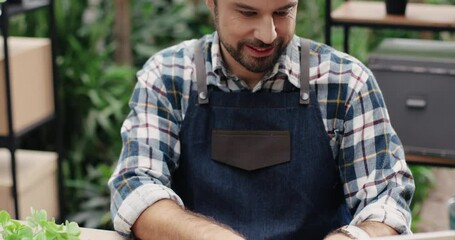 Close up of handsome cheerful Caucasian male florist in apron sitting at desk in floral store and typing on laptop. Joyful young man working and browsing on computer at workplace. Own business concept