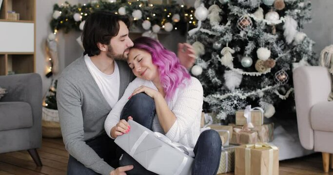 Happy couple in love celebrating Christmas together. Young man and woman are sitting together near Christmas tree cuddling and kissing each other. Husband and wife in decorated interior at home.