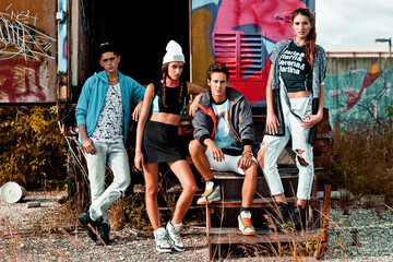 A portrait of an interracial group of 4 young adults lounging on wooden steps in front of an open graffiti sprayed container on a vacant lot during a pleasant summer day wearing cool urban outfits.