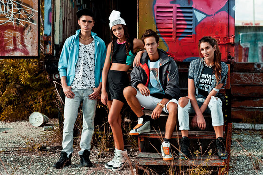 Portrait Of An Interracial Group Of 4 Young Adults Hanging Out On Wooden Steps In Front Of An Open Graffiti Sprayed Container On A Vacant Lot During A Warm Summer Day Wearing Cool Urban Outfits.