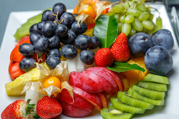 Assorted sliced fruit, black and white grapes, kiwi, prunes, pineapple, strawberries and physalis are on a platter. The view from the top.