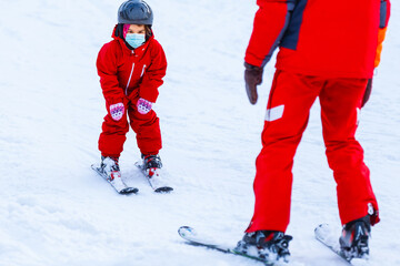 Portrait of a little girl skier in medical mask during COVID-19 coronavirus on a snowy mountain at...