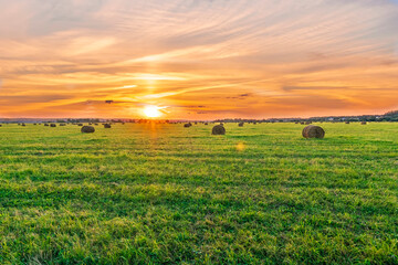 Obraz premium Scenic view at beautiful sunset in a green shiny field with hay stacks, cloudy sky, golden sun rays, anazing summer valley evening landscape