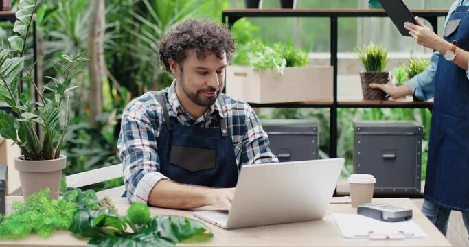 Portrait of happy Caucasian male and female florists in aprons working at flower shop and doing inventory. Man typing on laptop at desk in greenhouse. Woman tapping on tablet. Family business concept