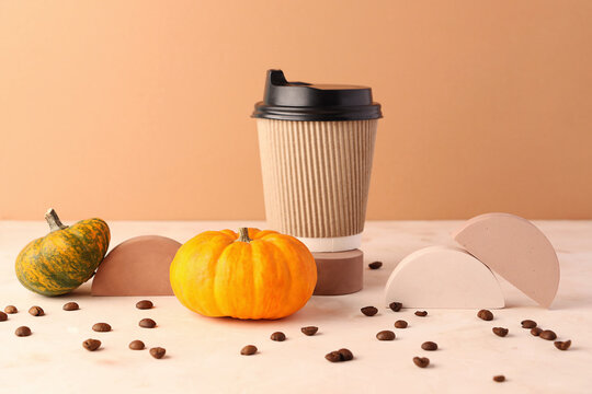 Craft paper coffee cup stand on the round podium.Empty geometric pedestals around it.Spicy mini pumpkins and coffee beans.Contemporary still life,seasonal latte concept.