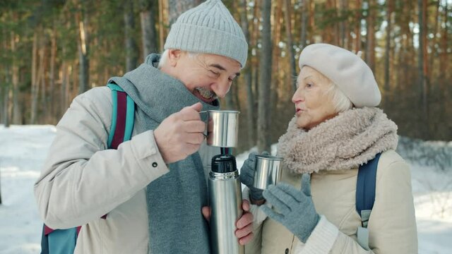 Happy senior couple man and woman are clinking cups drinking hot tea from thermos and chatting outdoors in winter park. Family leisure activities and tourism concept.