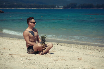 happy shirtless young hipster man  in shorts and sunglasses  with tattoo lying and holding pineapple on sea beach. Summer vacation. Tropical seascape with boats on the background..