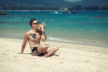 Stylish muscular hipster young man with tattoo wearing sunglasses trying to eat pineapple and sitting on the beach. Summer time concept. Phuket. Thailand.