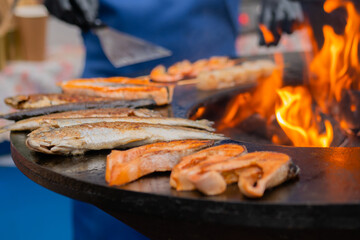 Process of grilling salmon steaks and mackerel fish on black brazier with hot flame at summer local food market - close up. Outdoor cooking, barbecue, gastronomy, seafood, cookery, street food concept