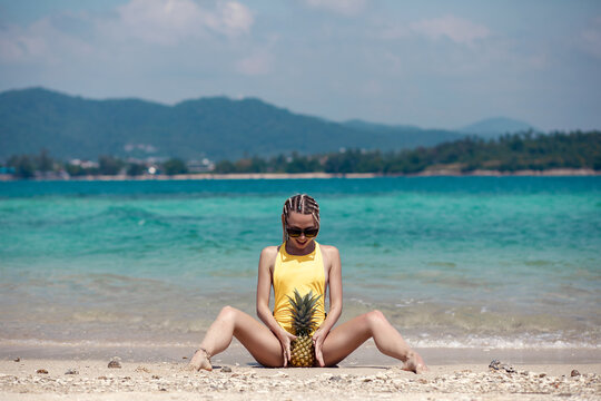 Sexy blonde girl with white dreadlocks  in bright yellow swimsuit sitting on the sand and holding pineapple between  her legs. tropical seascape. Summer vacation. Phuket. Thailand