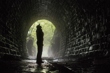Female silhouette with headlamp in wet brick tunnel, green forest background