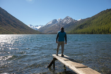 Young hiker wearing blue jacket standing by the mountain lake and looking at the distance