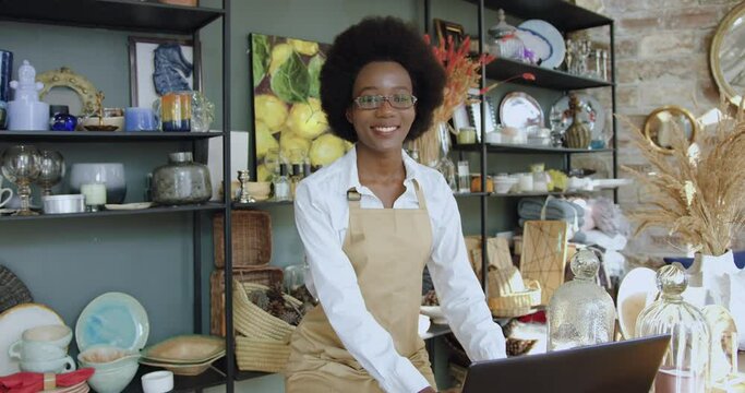 Adorable contented smiling young black-skinned woman with african hairstyle in apron poses on camera during working in beautiful souvenirs shop