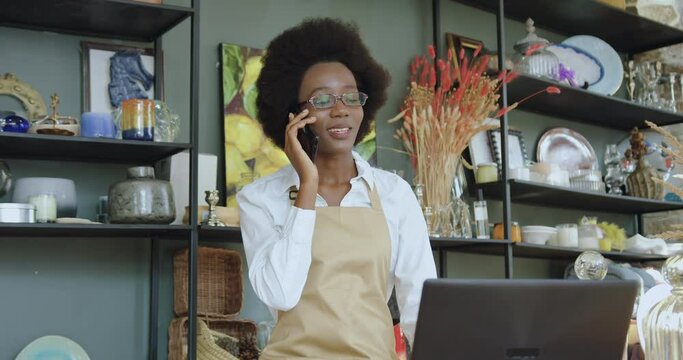 Portrait of good-natured happy smiling black-skinned woman with african hairstyle which talking on mobile while working in beautiful gift shop