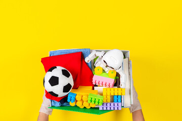 Donation concept. Woman hands with gloves holding donate box with clothes, books and toys on yellow background. Top view
