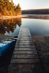 Fototapeta premium Moody mole (pier) and old blue boat in fall with crystal blue water and forest on background. Sunrise - mole (jetty) on the lake with water reflection of forest.