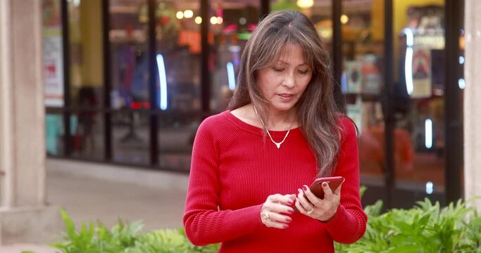 Face of beautiful exotic asian older woman with long dark hair wearing red long-sleeve top texting in front of glassed store front. Medium shot of her looking down. Bokeh background of glass windows.