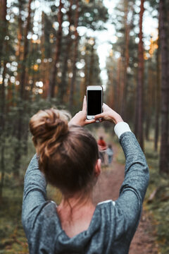 Young woman taking photos using smartphone during walk in a forest on summer vacation trip. Back view of girl holding smartphone
