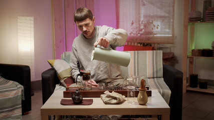 A young man prepares for a romantic date with an authentic japanese tea ceremony at home, pours boiling water into a teapot with an infuser.