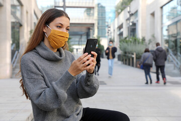 Attractive young business woman with face mask sitting on bench of pedestrian city street using a smartphone