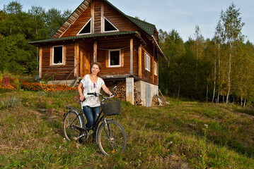 Woman with a bicycle in summer. Bike ride near the wooden cottage. Flowers, greens and forest in the background.
