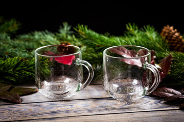 Two empty glass transparent cups or mugs on a vintage wooden glass table decorated by branches of christmas tree and holly leaves on black background. Cups for christmas hot drinks.