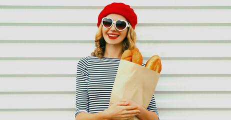 Close up of laughing young woman wearing a french red beret holding paper bag with long white bread baguette over a white background