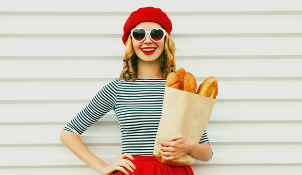 Close up of laughing young woman wearing a french red beret holding paper bag with long white bread baguette over a white background