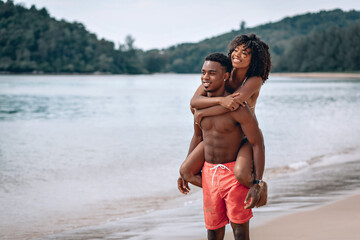 Young African man carrying his smiling girlfriend on his back while enjoying a late afternoon together at the beach. Phuket. Thailand.