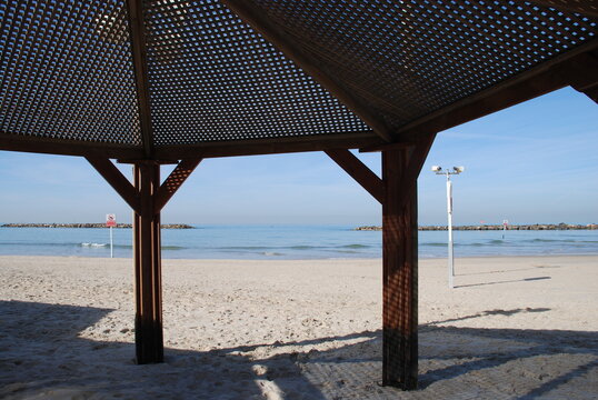 Sea view from under a wooden shed.
Mediterranean sea in winter. Small waves are splashing along the coast. Wooden canopy with mesh roof. Blue sky, pillars dug into the ground.
