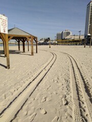Car tracks on the beach sand.
Beach sand on the sand footprints of a person and a footprint from a car. There are wooden awnings from the sun. In the distance water and city buildings.