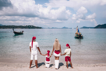 Beautiful family in christmas costumes and santa hat stand on the beach, looking at the sea where a small boat floats. Christmas and New Year concept. Vacation. Phuket. Thailand