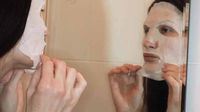 Woman in white fabric regenerating collagen cosmetic mask looking in mirror, enjoy at home natural beauty and healthy care treatment