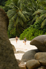 Young children walk together under palm trees, at tropcial sandy beach behind huge rocks