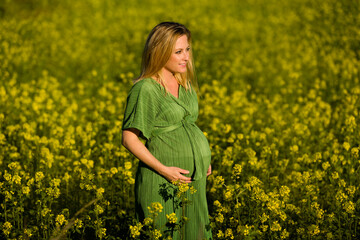 Beautiful and happy blonde expecting mother in green dress, smiles in the middle of a yellow flowery field.