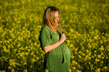 Beautiful and happy blonde expecting mother in green dress, smiles in the middle of a yellow flowery field.