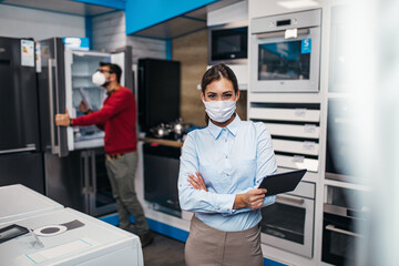 Young saleswoman working in store with household appliances. She is wearing face protective mask and holding tablet. Pandemia, Covid-19 concept.