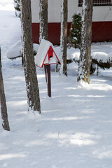 A small red and white birdhouse on a stand that is covered and surrounded by snow, in a yard between trees.Copy space, front view, empty space for text.