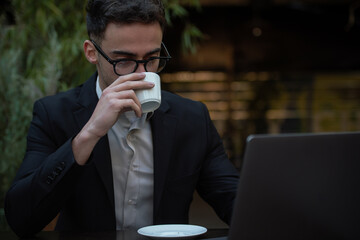 Young successful businessman working on a laptop while sitting in coffee bar during work break lunch. Speaking on his phone while working and drinking coffee.	