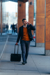 Young businessman walking through a station while talking on the phone and pulling his travel bag.