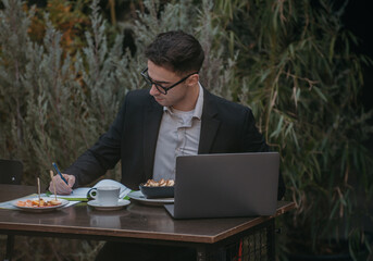 Young successful businessman working on a laptop while sitting in coffee bar during work break lunch. Speaking on his phone while working and drinking coffee.	