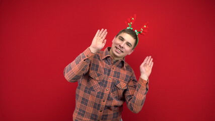 A young man is dancing with a headband in the form of Christmas antlers. Studio shooting on a red background.