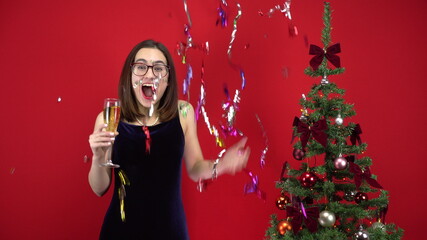 Young woman counts five seconds to the holiday with a glass of champagne near the Christmas tree on a red background. A girl with glasses and an evening dress.