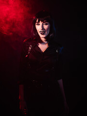 An image of a vampire woman in a black shirt, harness, black wig, her gaze directed towards the camera with a smile from her fangs.