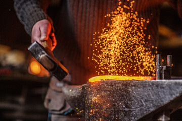 The blacksmith hits the red-hot workpiece in the forge with a hammer and glowing sparks fly in all...