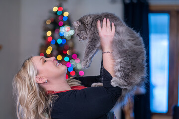Adorable blonde woman holds a grey cat, with Christmas tree bokeh in background