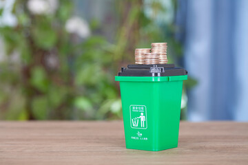 A model of a green garbage can with food waste and a pile of dollar coins on its lid