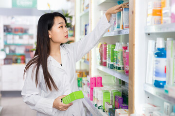 Glad positive chinese woman pharmacist keeps track of drugs in interior of pharmacy